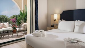 Hotelkamer met een bed, nachtkastje, lamp, raam met uitzicht op groen, en een balkon met tafel en stoelen.