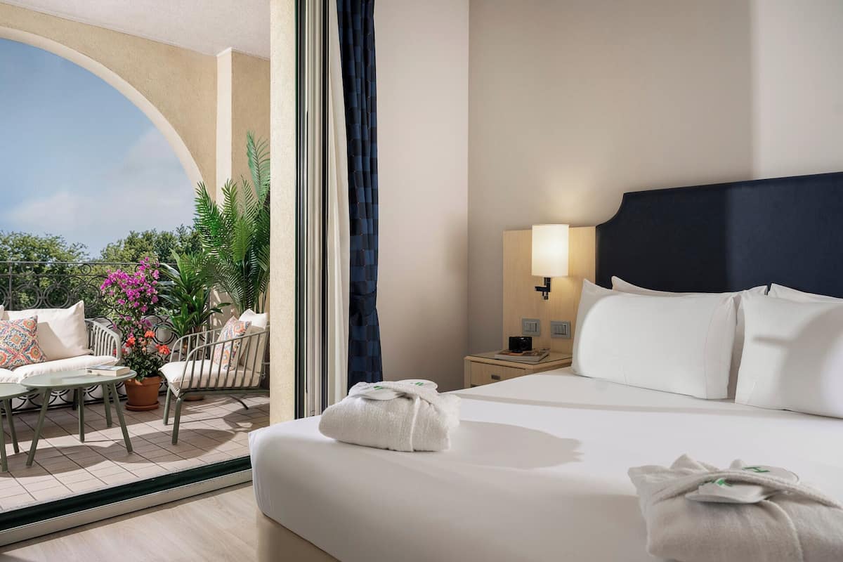 A hotel room with a bed, a nightstand, a lamp, a window with a view of greenery, and a balcony with a table and chairs.