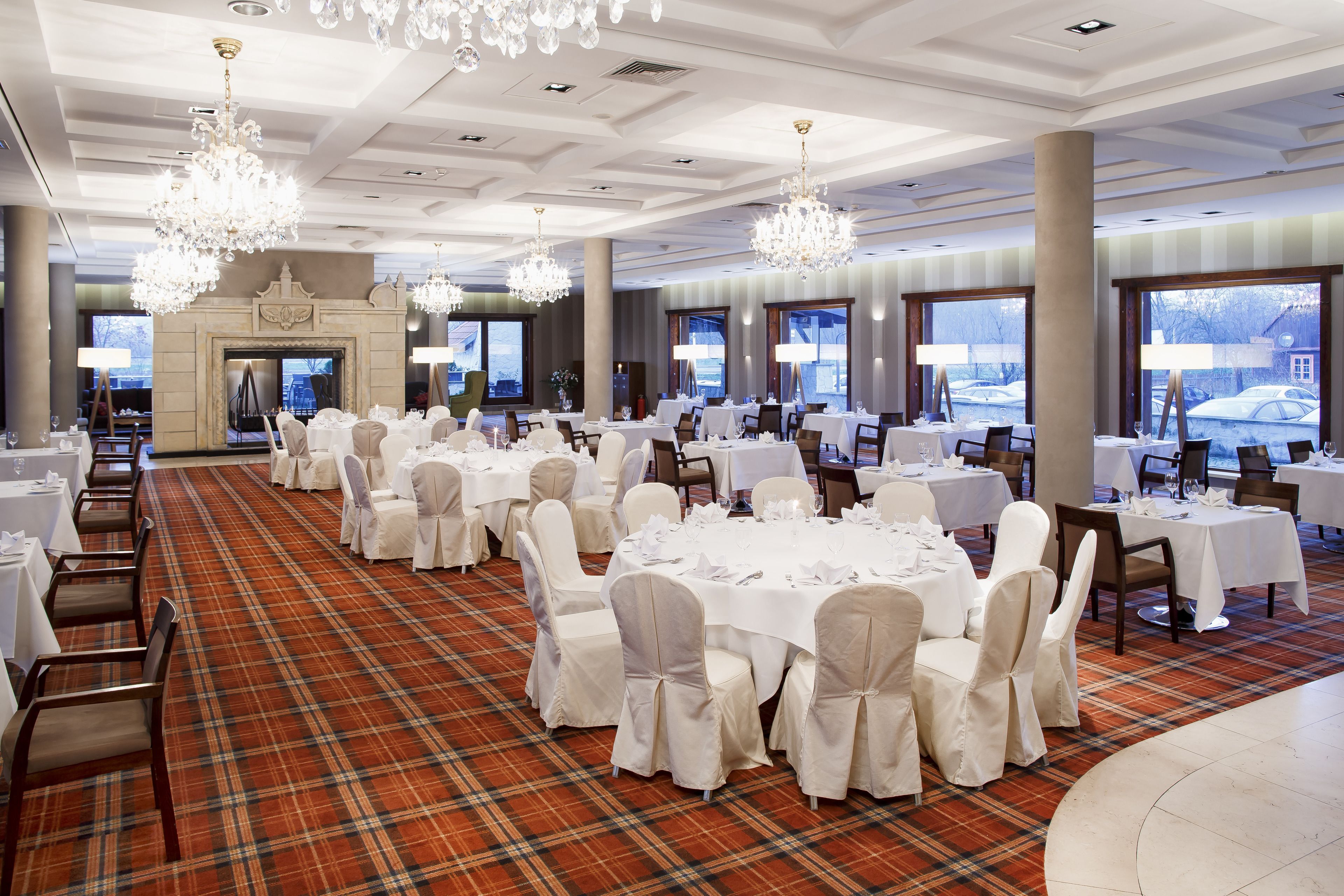 A banquet hall with round tables set for dining, white chairs, chandeliers, and large windows with a view.