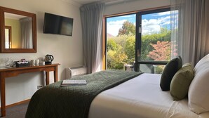 A hotel room with a bed, a desk with a flat-screen TV, a coffee maker, a thermostat, and a window with a view of trees. - Wanaka Springs Lodge (Wanaka)