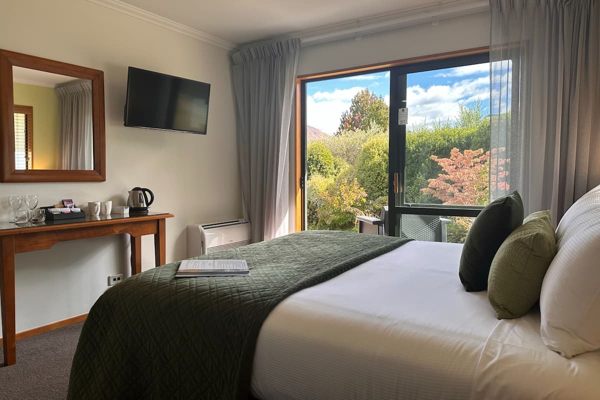 A hotel room with a bed, a desk with a flat-screen TV, a coffee maker, a thermostat, and a window with a view of trees.