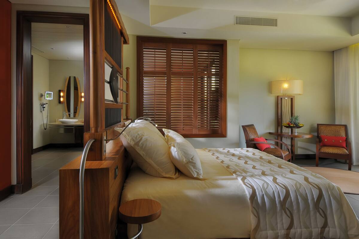 A hotel room with a wooden bed, a bedside table, a chair, a desk with a lamp, and a window with blinds.