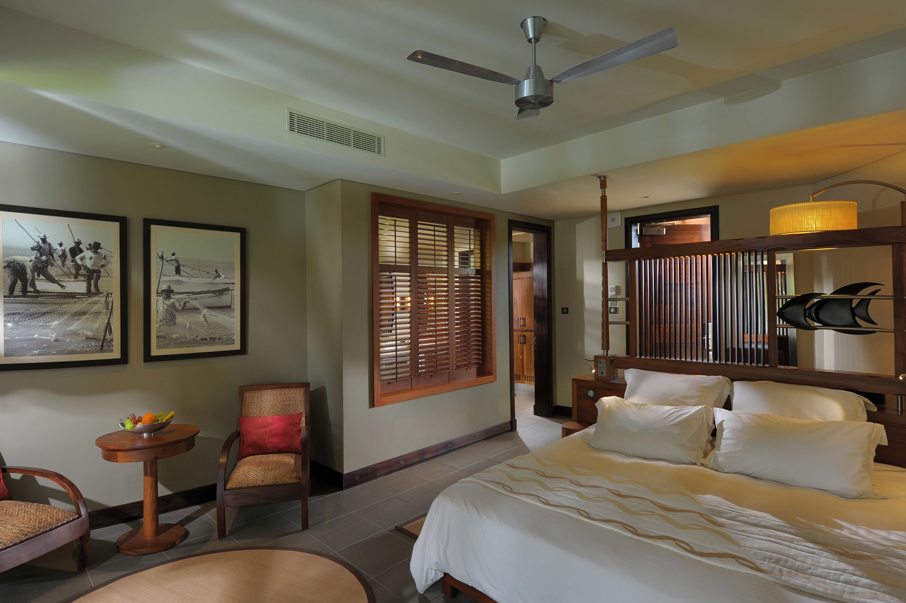 Habitación de hotel con cama, una silla, una mesita con fruta, ventilador de techo y contraventanas de madera.