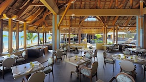 A spacious outdoor dining area with wooden beams and thatched roof, overlooking a beach. Tables are set with tableware and drinks. - Trou aux Biches Beachcomber Golf Resort & Spa (Trou aux Biches)