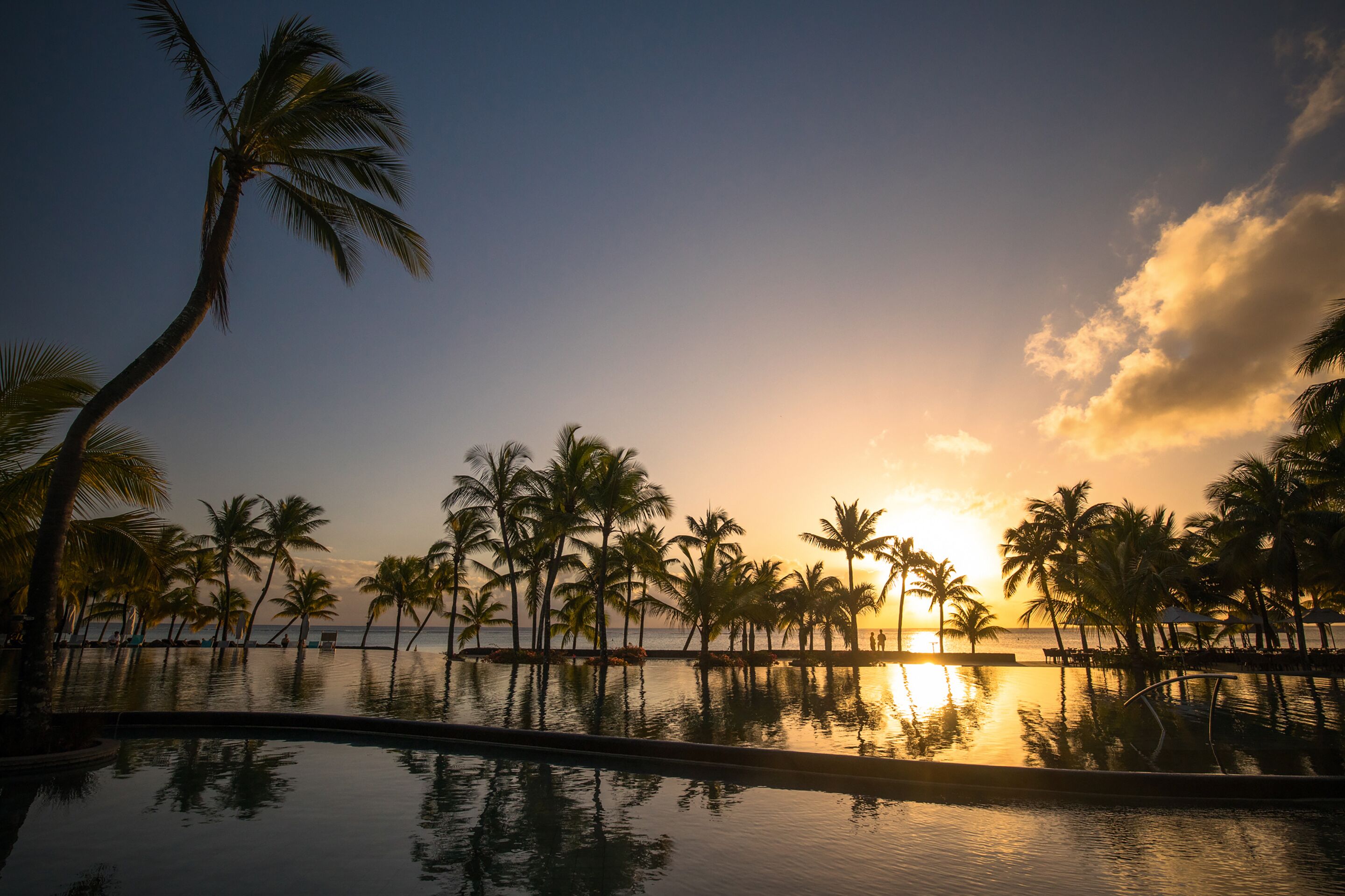 A sunset view over a pool with palm trees and a beach in the background.