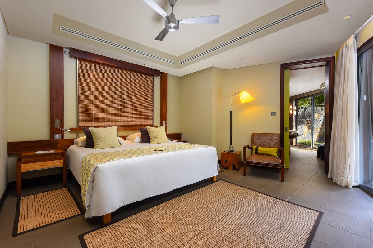 A hotel room with a large bed, a bedside table, a chair, a standing lamp, and a view of the outdoors.