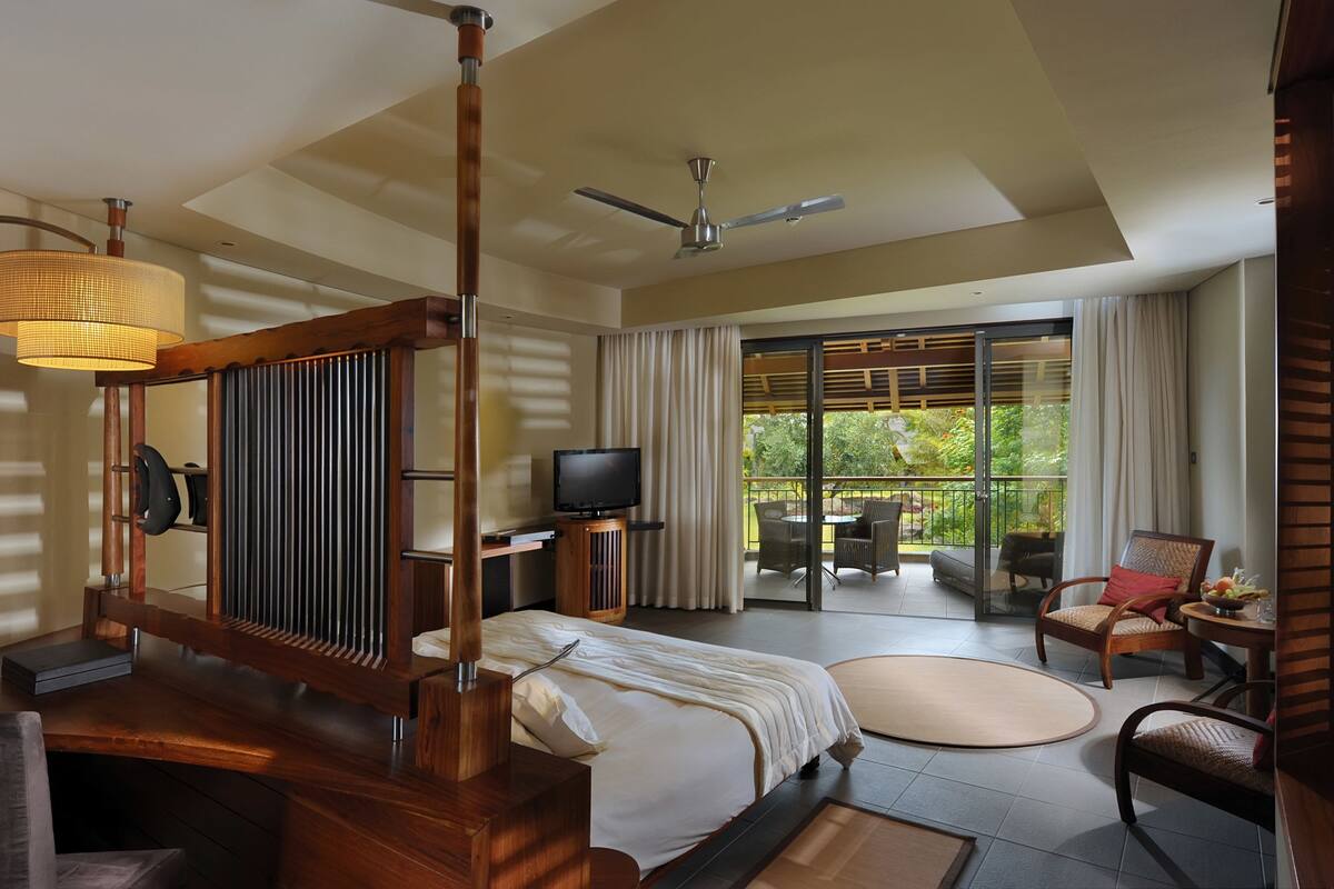 A modern hotel room with a large bed, a television, a balcony with outdoor furniture, and a ceiling fan.