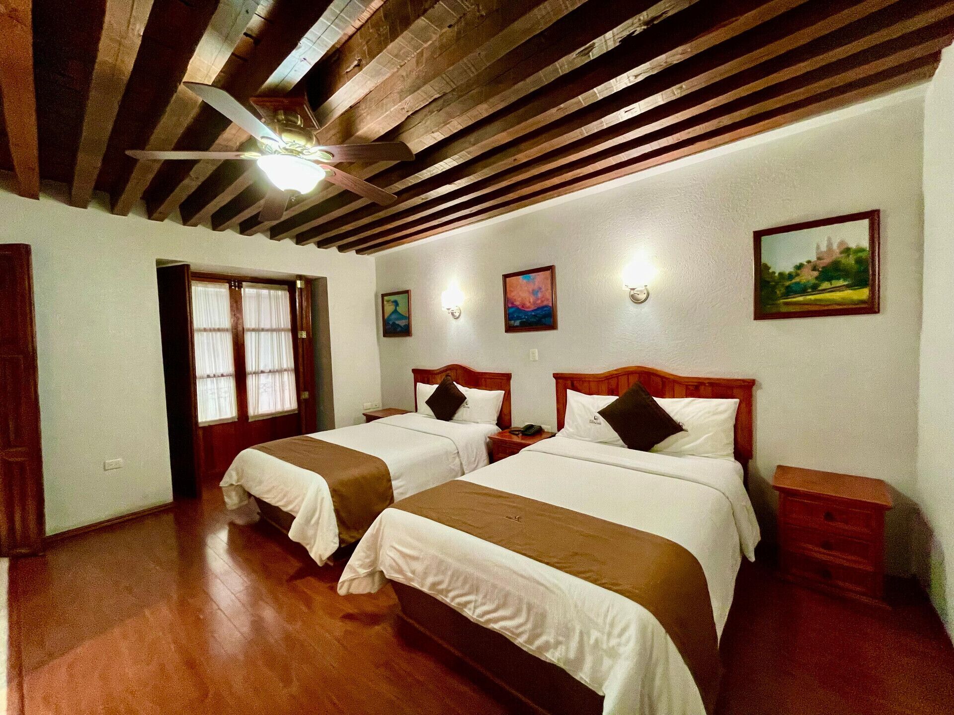 A hotel room with two beds, wooden beams on the ceiling, a window with curtains, and wall-mounted artwork.