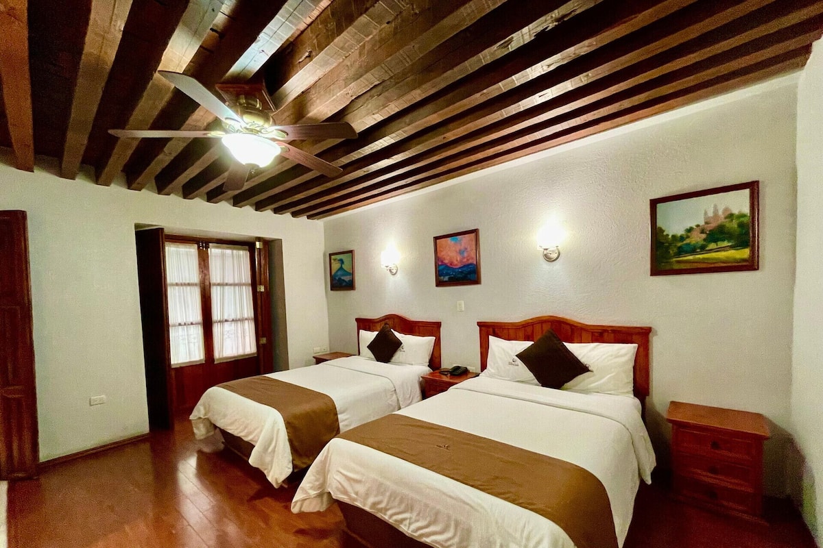 A hotel room with two beds, wooden beams on the ceiling, a window with curtains, and wall-mounted artwork.