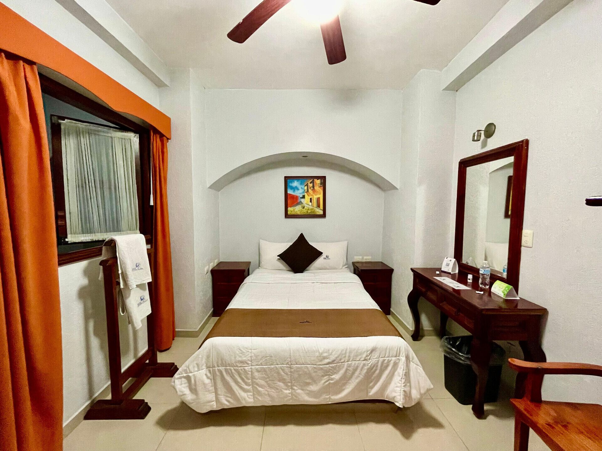 A hotel room with a bed, a desk, a chair, a mirror, and a painting on the wall.