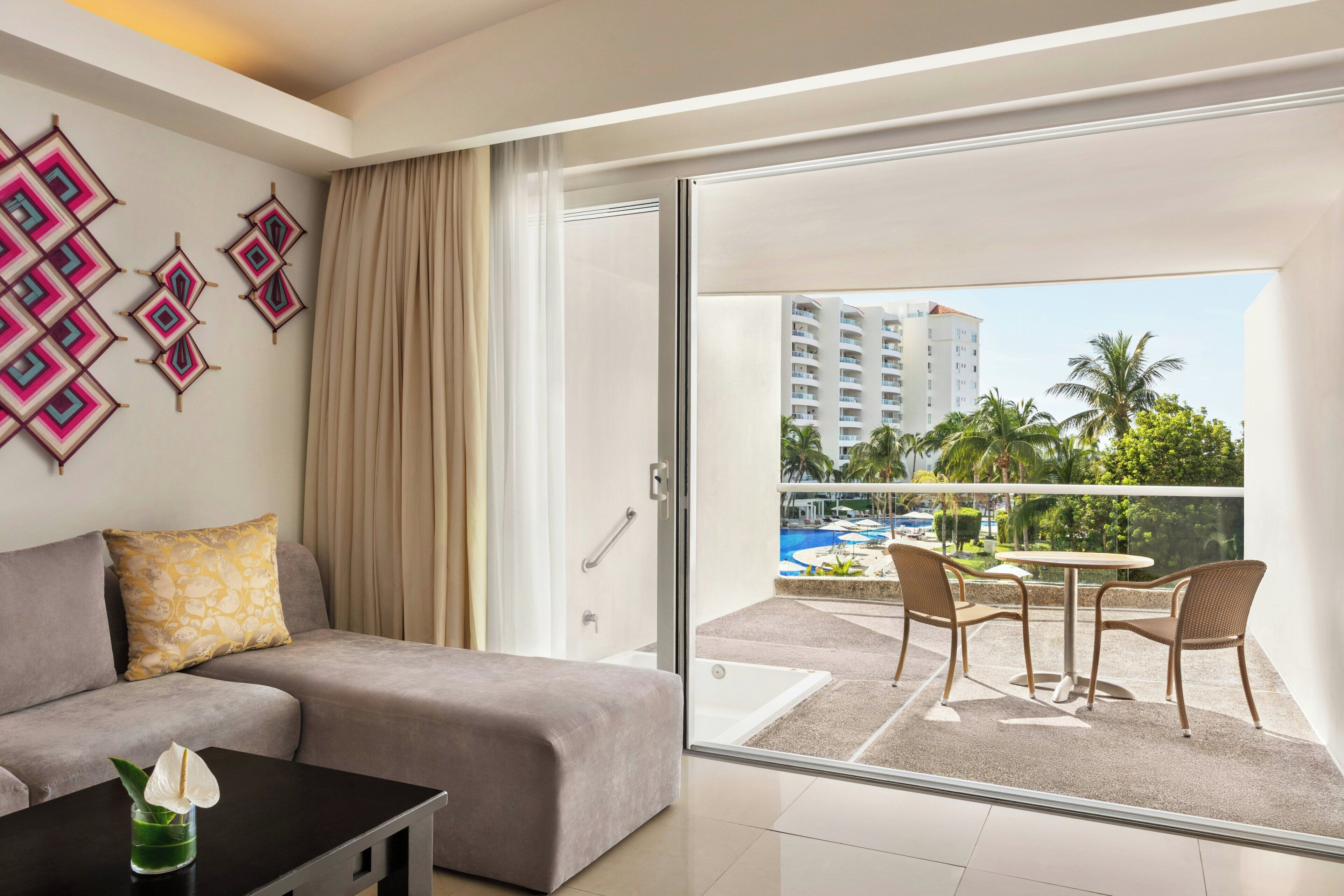 Habitación de hotel moderna con balcón, sofá, mesa de centro y vista a una piscina y edificios.