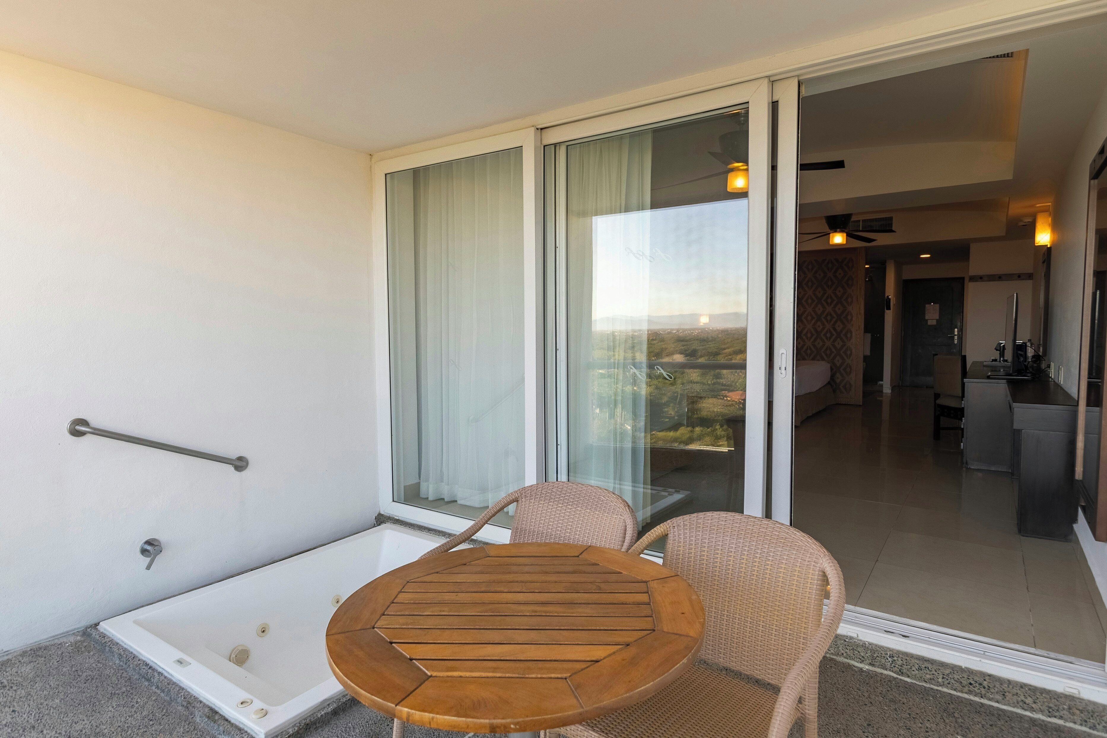 A balcony with a glass door, a wooden table, two wicker chairs, and a view of a cityscape.