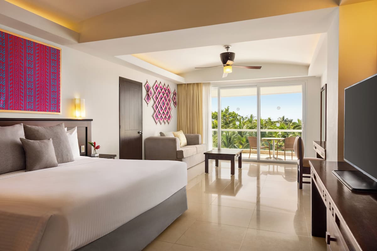 Une chambre d’hôtel avec un grand lit, un canapé, une télévision, un ventilateur de plafond et un balcon donnant sur un paysage verdoyant.