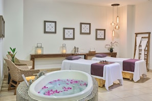 Une salle de spa dotée d’une grande baignoire remplie d’eau et de pétales de rose, de deux tables de massage recouvertes de draps blancs, de chaises en osier et d’œuvres d’art décoratives murales.