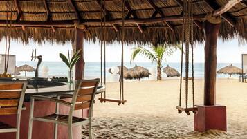 Ein Restaurant direkt am Strand mit einem Reetdach, Holzstühlen und Blick aufs Meer.