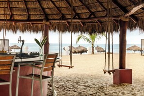 Un restaurante en la playa con techo de paja, sillas de madera y vista al océano.