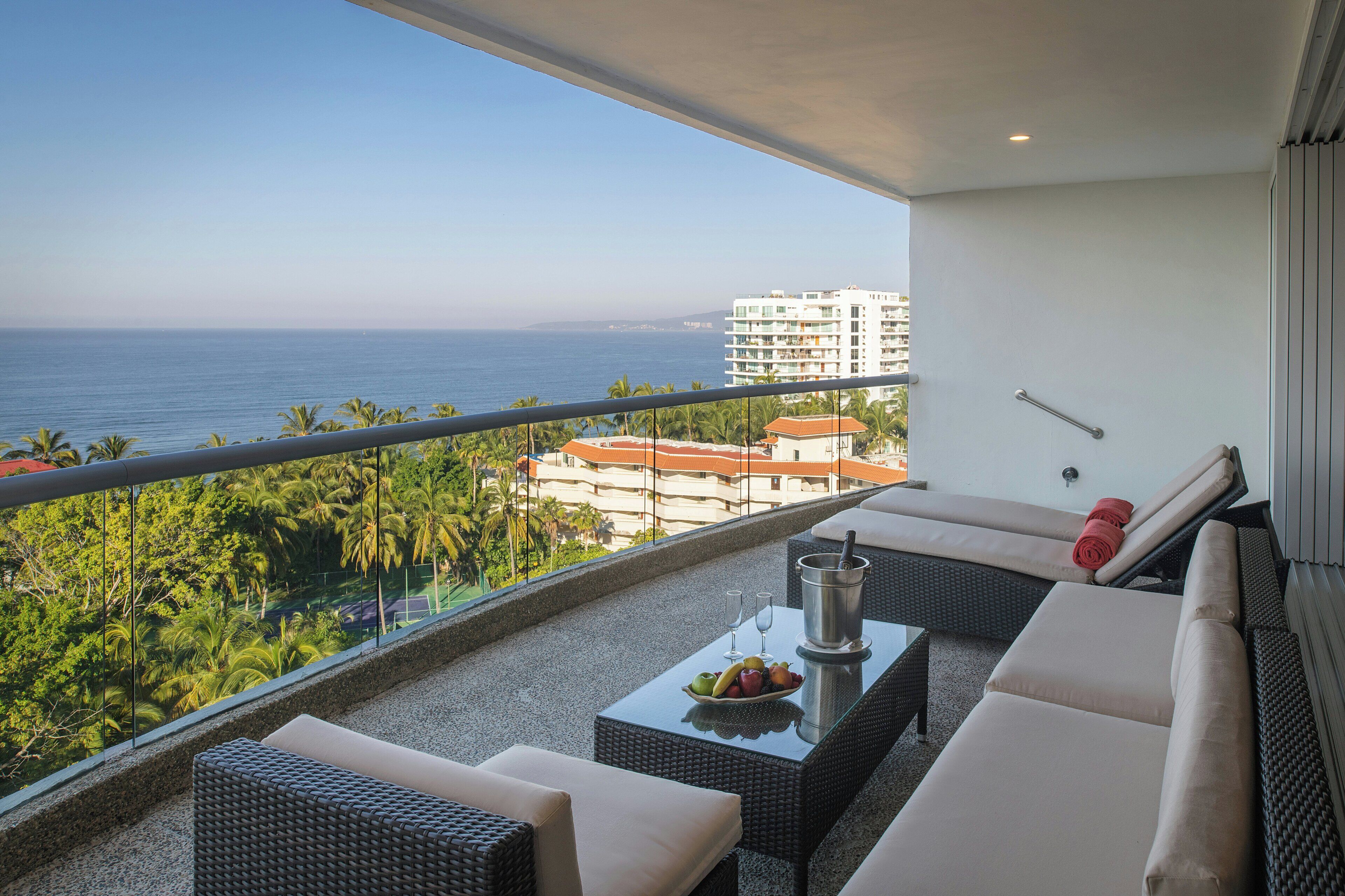 Un balcón con vista al mar, amueblado con un sofá, dos sillas y una mesa con un frutero y un recipiente metálico.