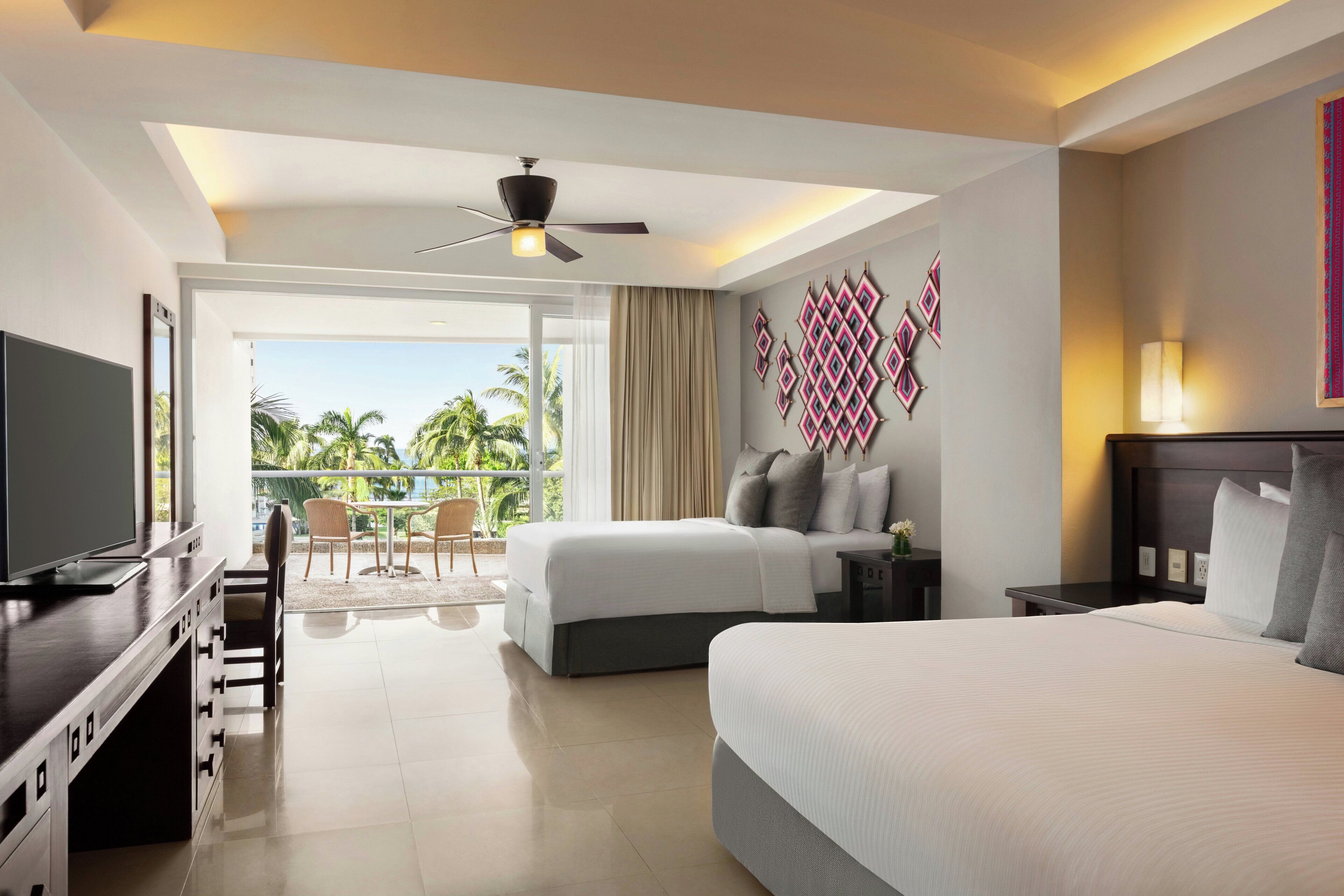A hotel room with a bed, a desk with a TV, a balcony with a view of palm trees, and a ceiling fan.