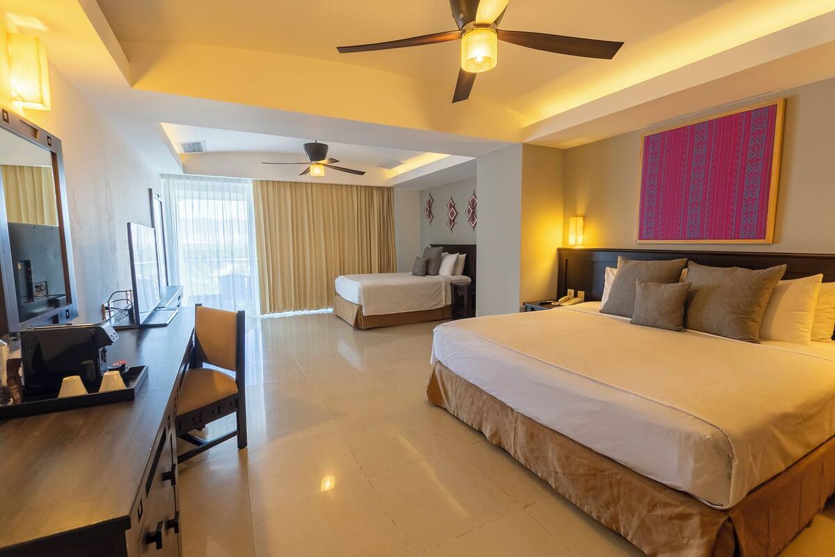 A hotel room with a large bed, a desk with a chair, a ceiling fan, and a large window with curtains.