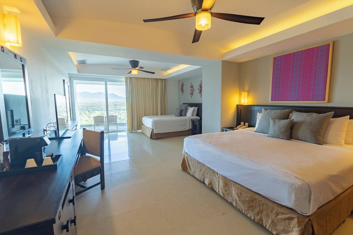 A hotel room with a large bed, a desk, a chair, a ceiling fan, and a large window with a view.
