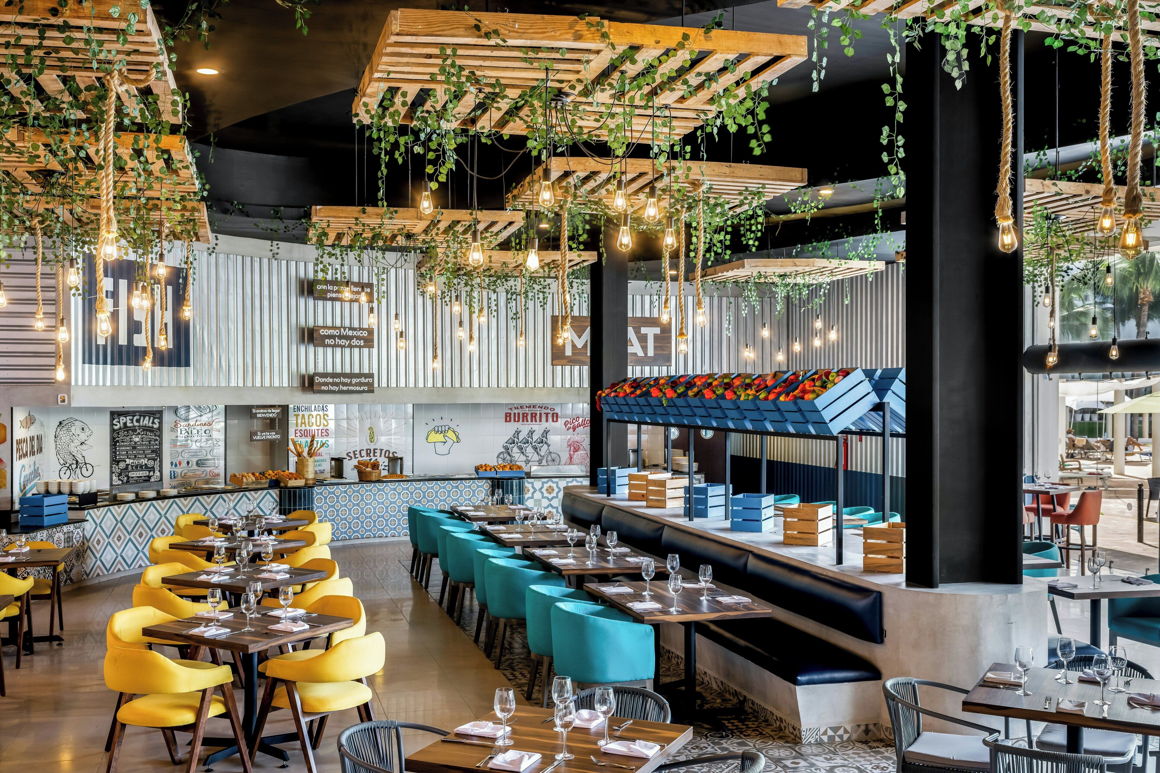 A restaurant interior with high ceilings, hanging plants, and exposed wooden beams. Tables are set with wine glasses and plates, and there are blue cushioned seating areas.
