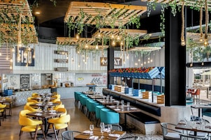 A restaurant interior with high ceilings, hanging plants, and exposed wooden beams. Tables are set with wine glasses and plates, and there are blue cushioned seating areas.