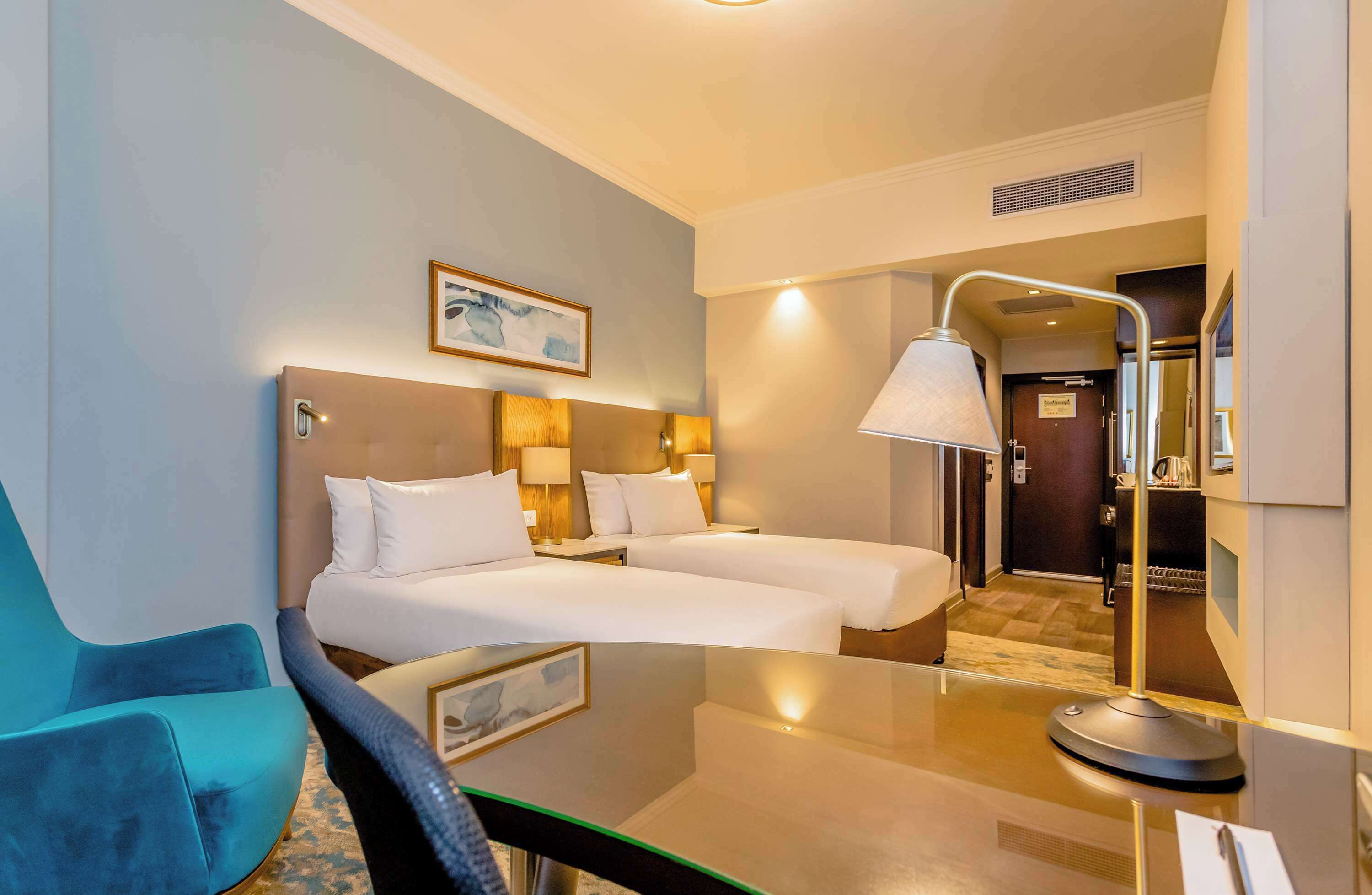A hotel room with two beds, a desk, a chair, a lamp, and a painting on the wall.