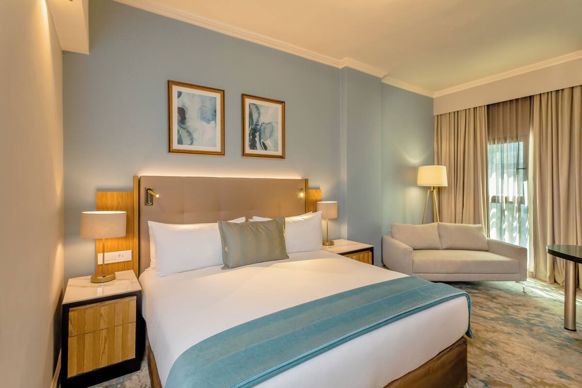 A hotel room with a large bed, two bedside tables with lamps, a sofa, and two framed artworks on the wall.