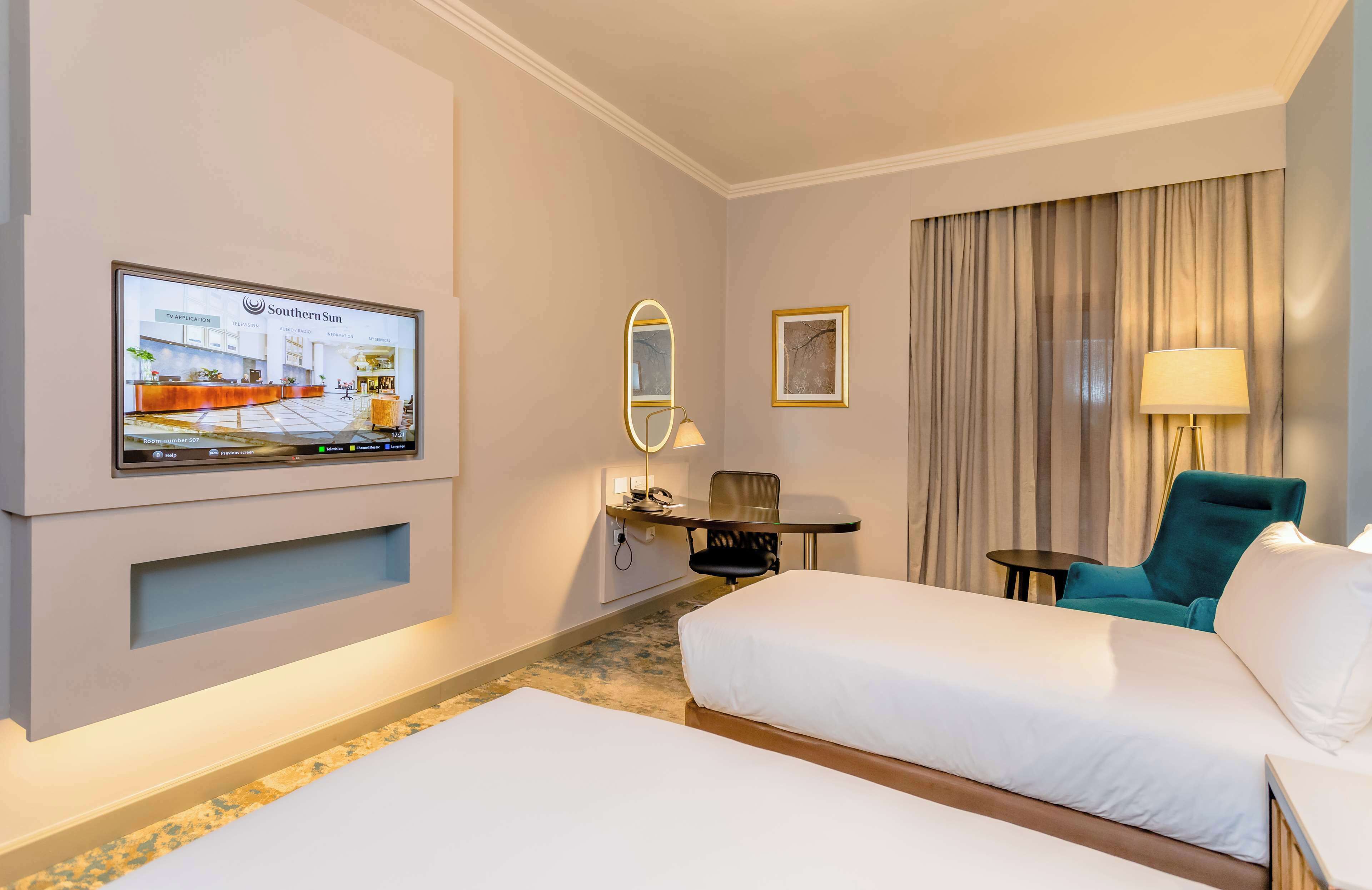 A hotel room with two beds, a TV mounted on the wall, a desk with a chair, a lamp, and a framed picture.