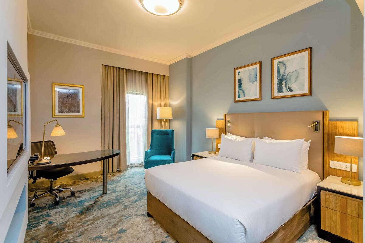 A hotel room with a large bed, a desk with a chair, a desk lamp, a bedside table with a lamp, and two framed pictures on the wall.