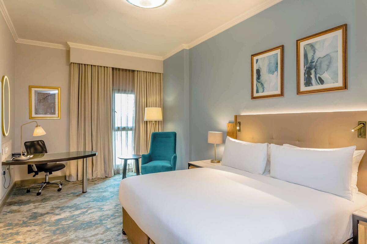 A hotel room with a large bed, a desk with a chair, a small table, and two framed artworks on the wall.