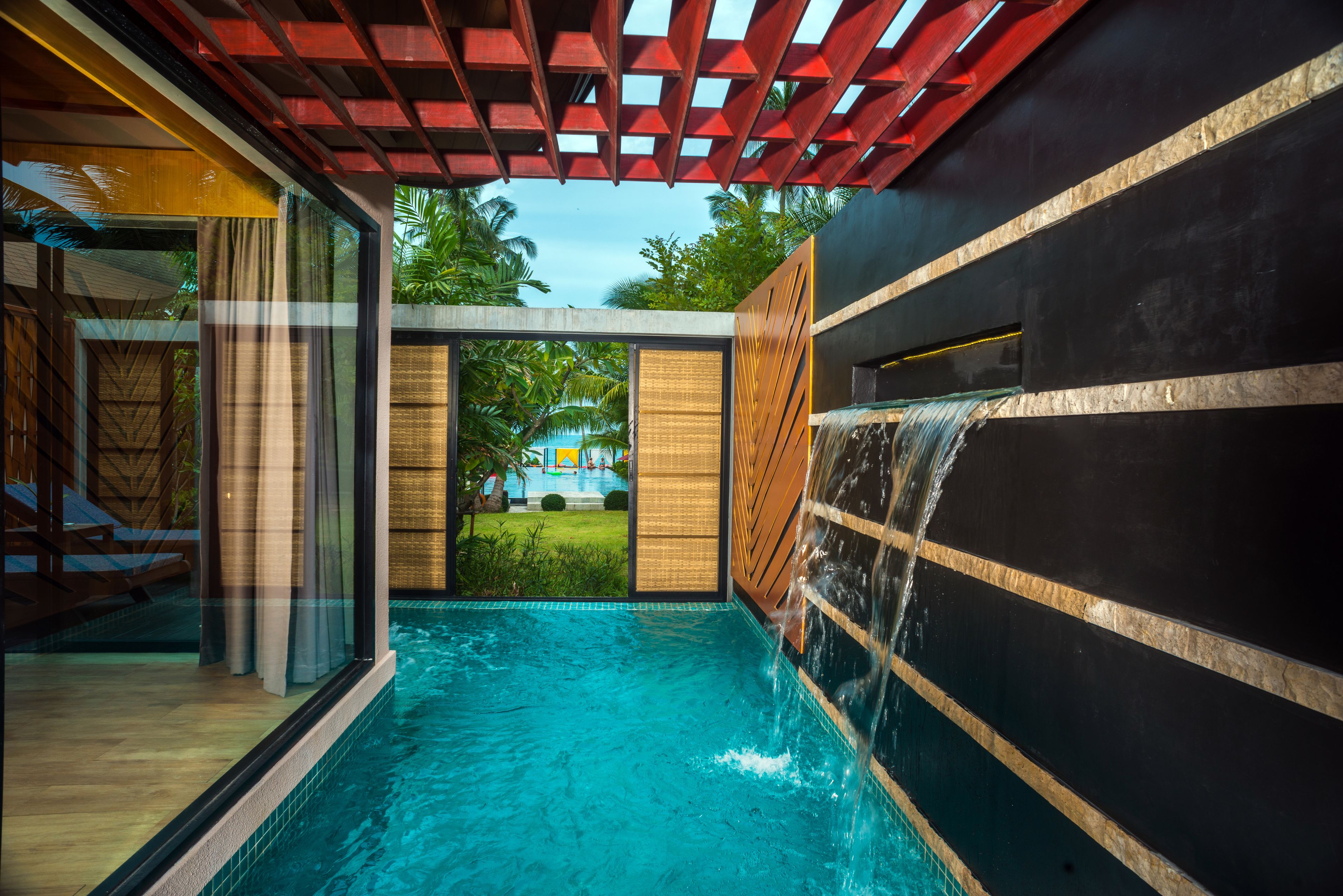 A modern indoor pool with a waterfall feature, glass walls, and a view of a tropical garden.