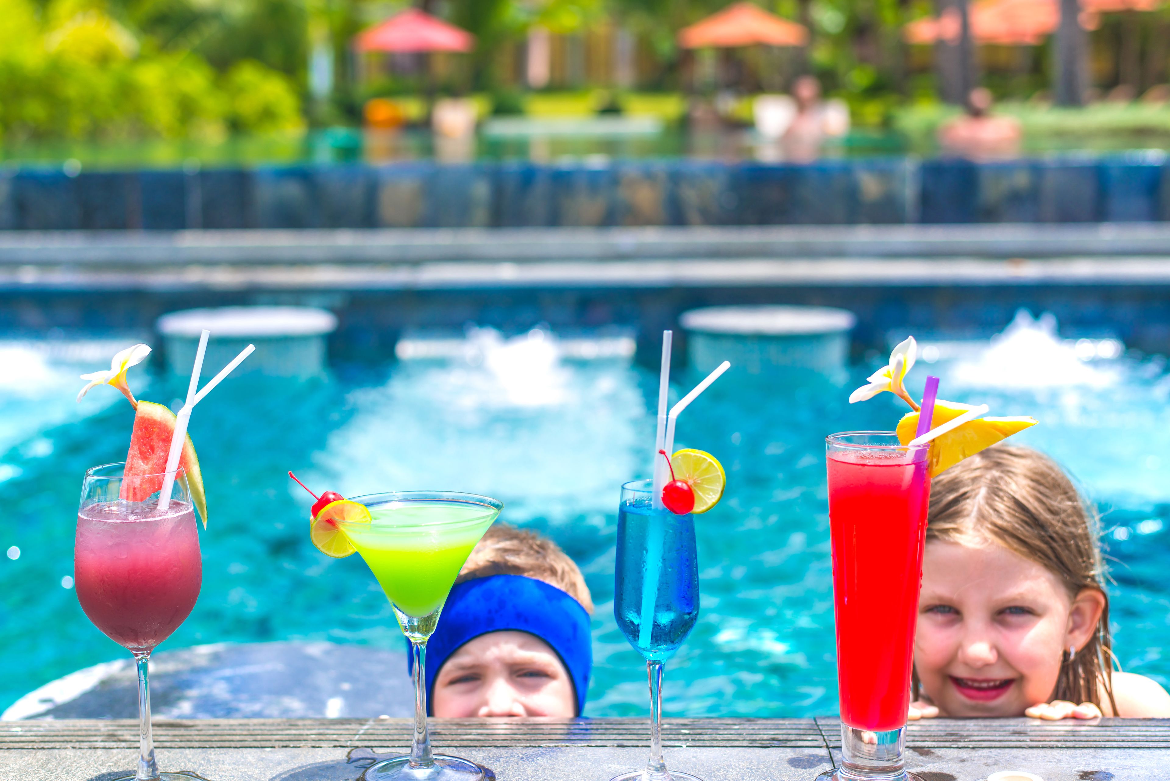A poolside setting with three drinks in glasses, each garnished with a fruit and a straw.