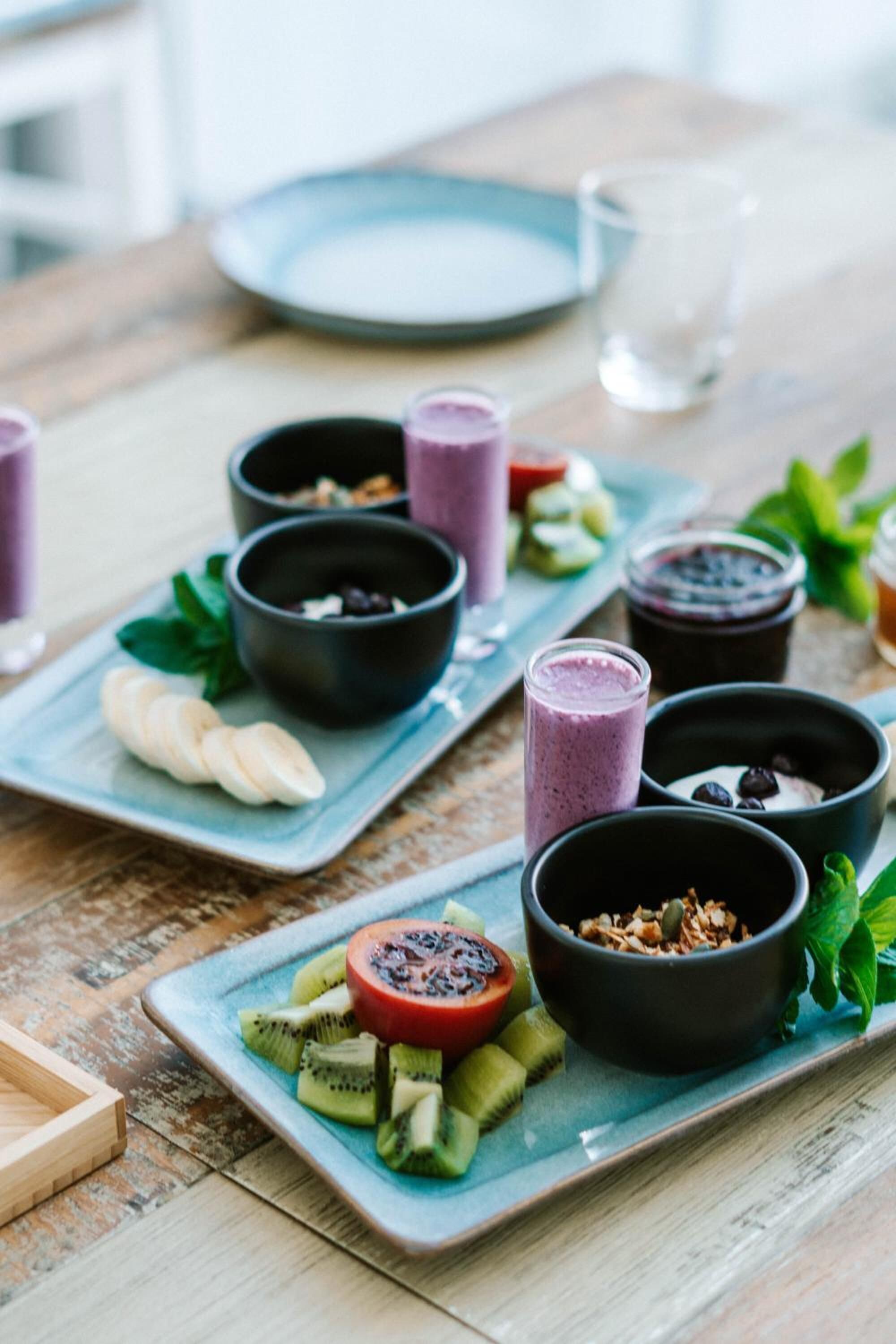 Una mesa de madera con dos platos cuadrados que contienen recipientes con comida y bebidas, y un vaso de agua.