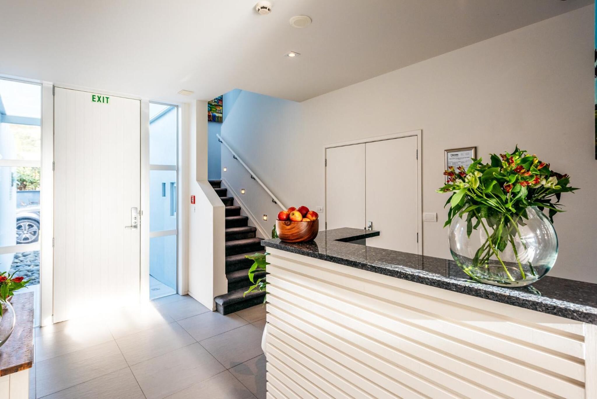 A modern reception area with a white reception desk, a glass vase with flowers, a fruit bowl, and a staircase leading to an upper floor.