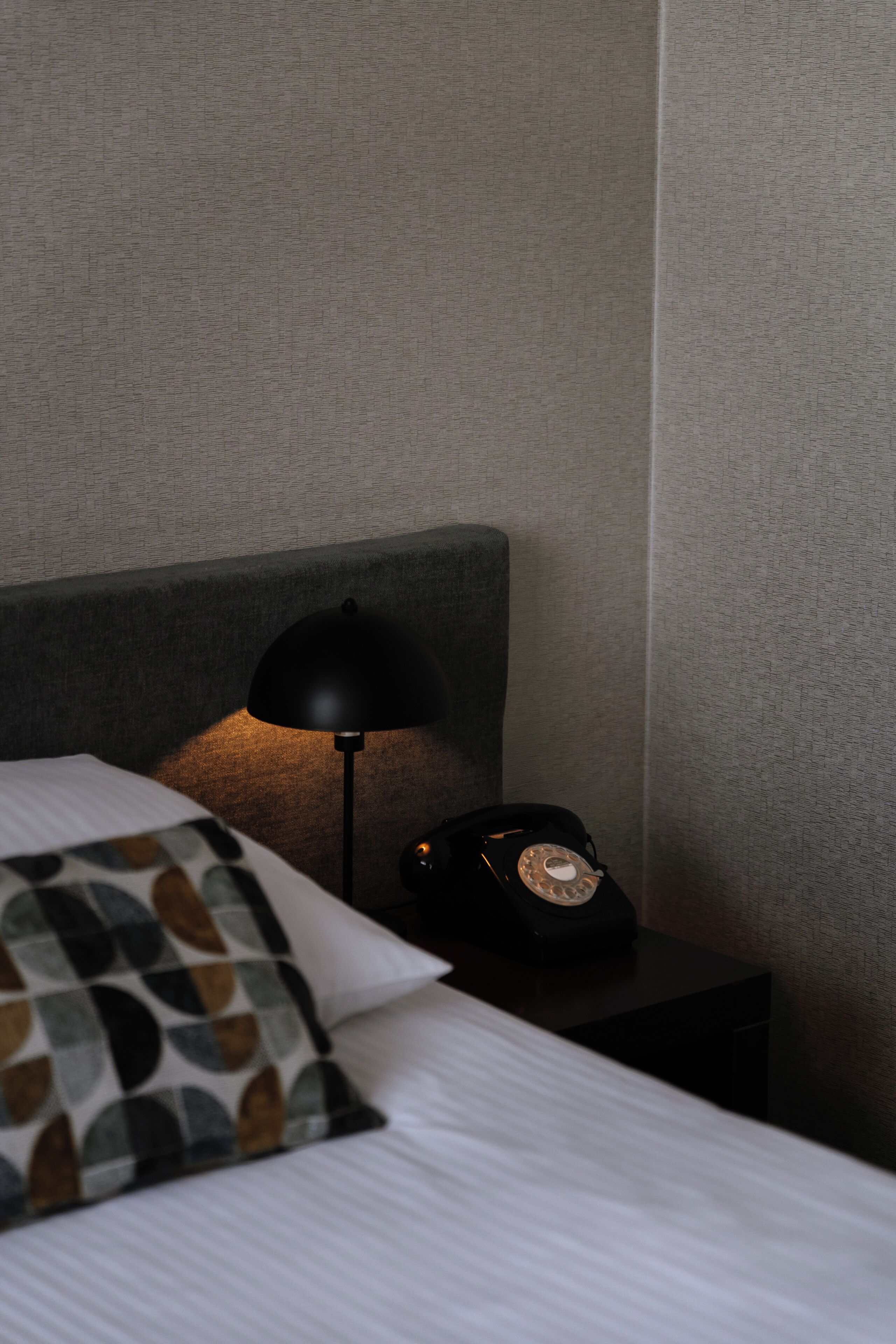 A neatly made bed with a patterned pillow, a black lamp, and a vintage telephone on a bedside table.