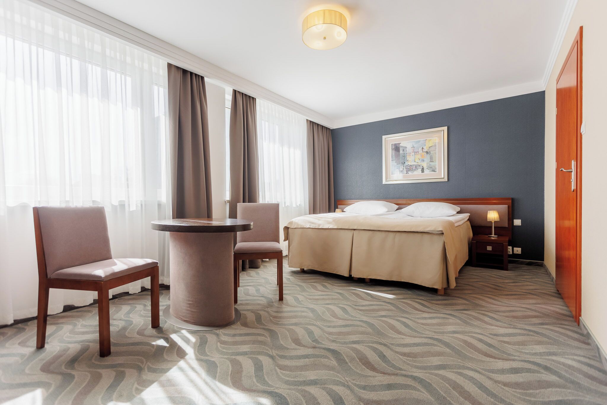 Una habitación de hotel con una cama, un escritorio, dos sillas, una lámpara y un cuadro en la pared.