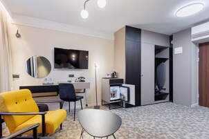 Quarto de hotel moderno com sofá, cadeira, escrivaninha, TV de tela plana, espelho, guarda-roupa e mini-geladeira.