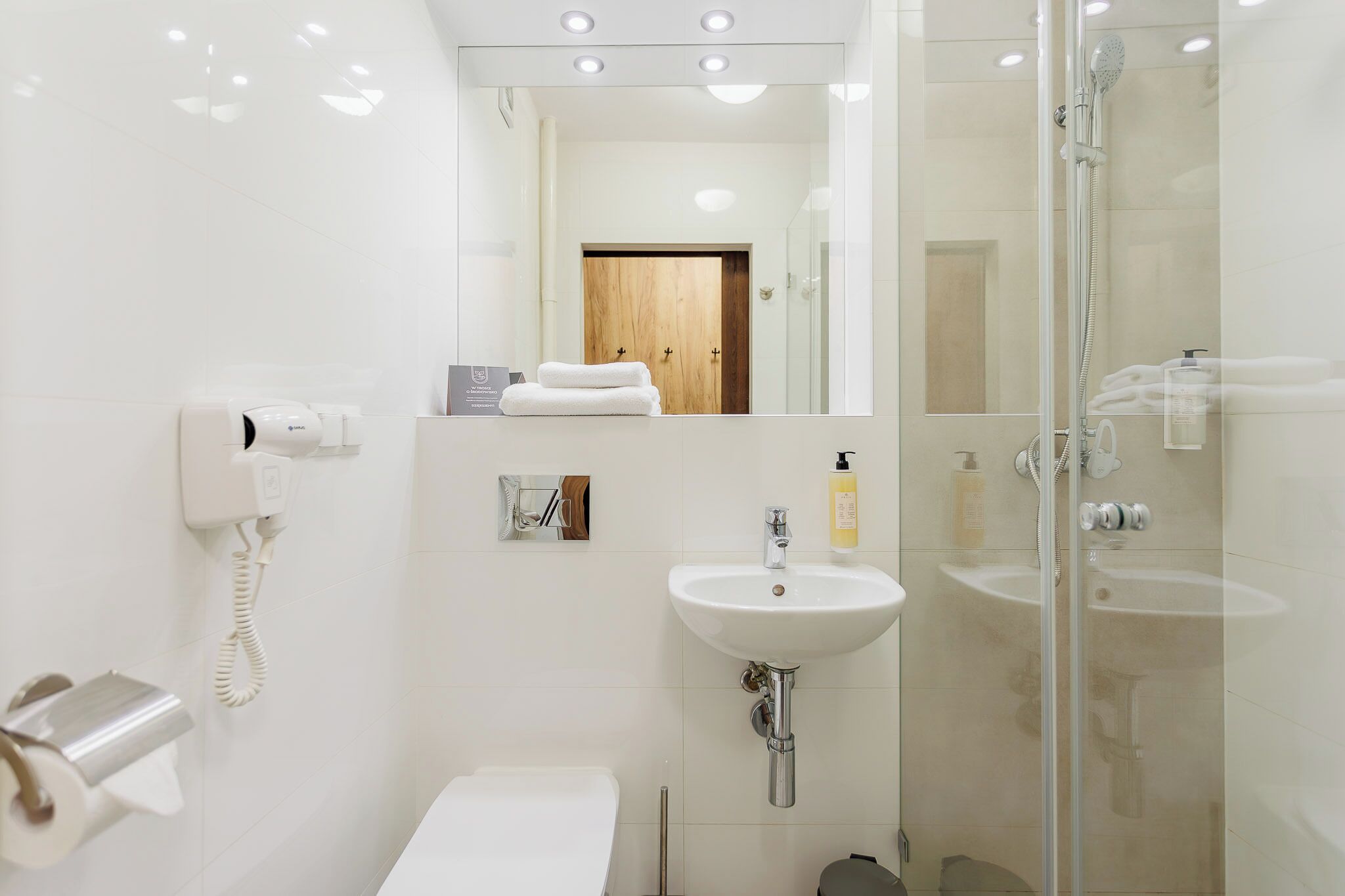 Un baño moderno con cabina de ducha de vidrio, un lavabo blanco y un inodoro. Hay un espejo sobre el lavabo, y un perchero con toallas.