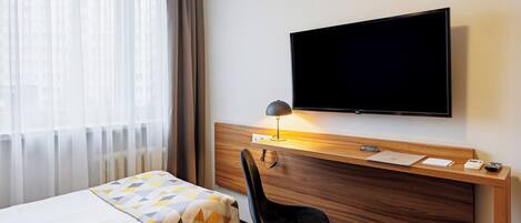 A hotel room with a bed, a desk with a flat-screen TV, a lamp, and a chair.