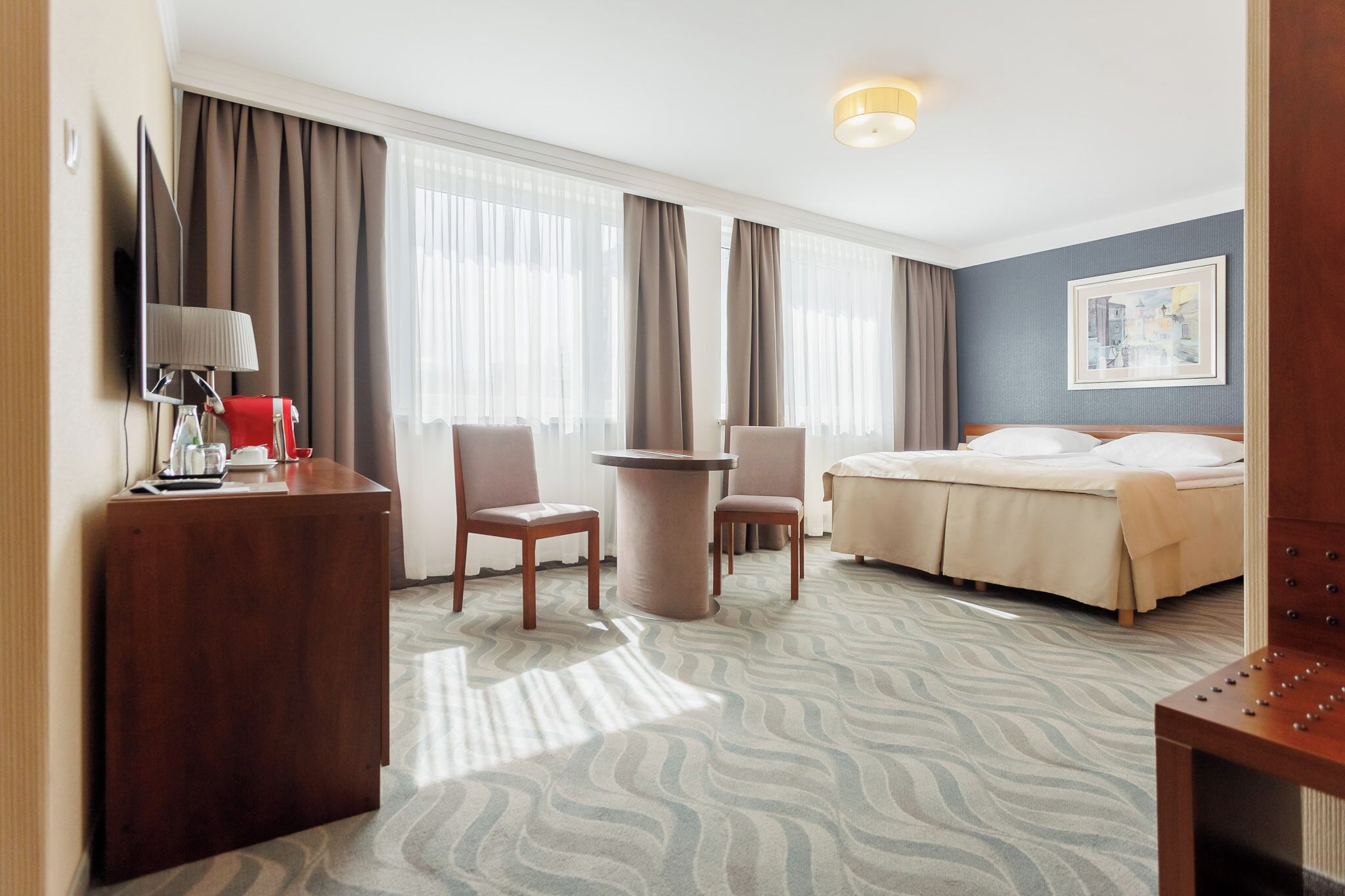 Et hotelværelse med en seng, to stole, et skrivebord med lampe, et fjernsyn og et stort vindue med gardiner.