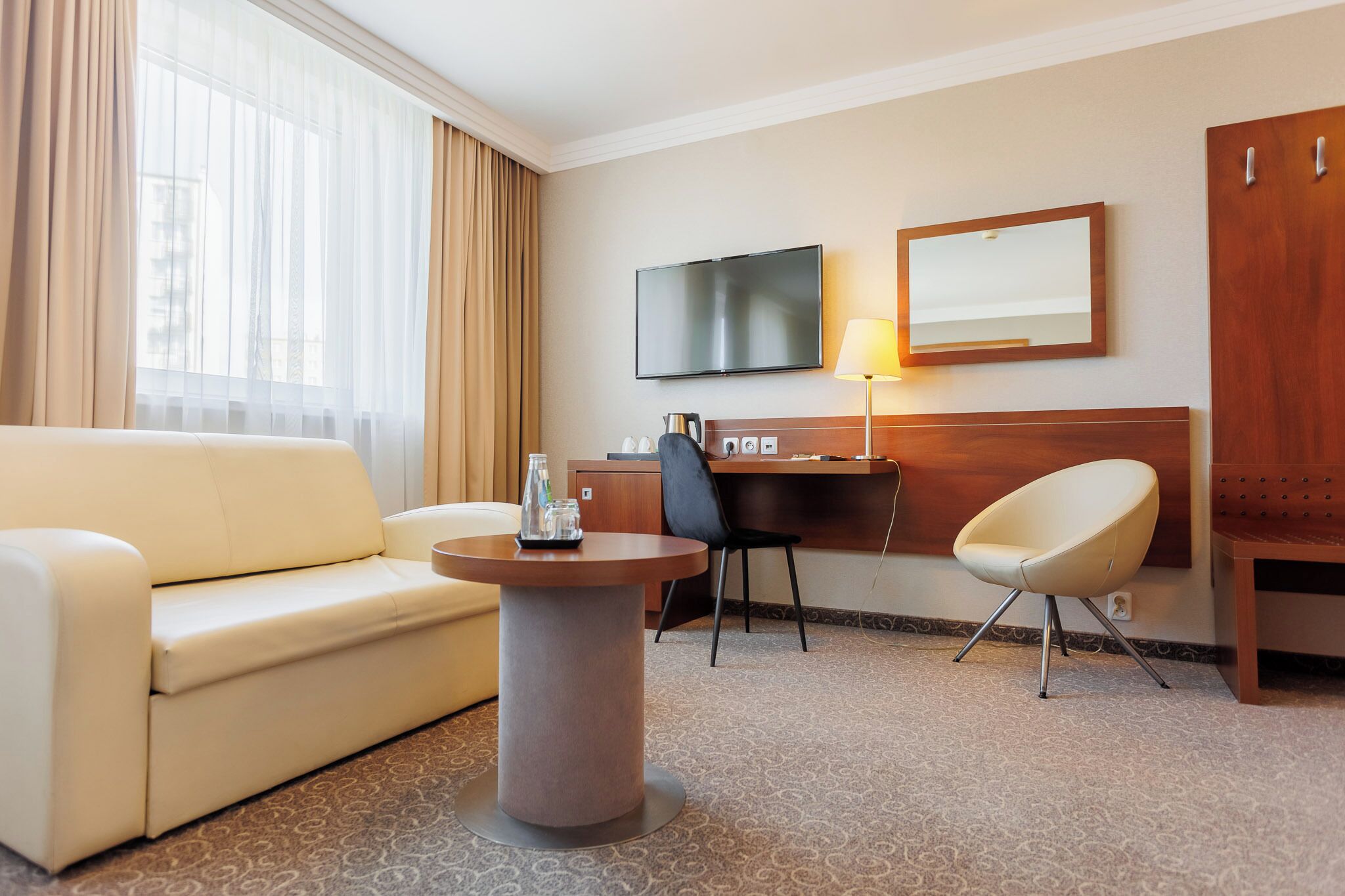 A hotel room with a sofa, a round coffee table, a desk with a chair, a television, a mirror, and a wardrobe.