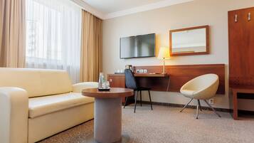 A hotel room with a sofa, a round coffee table, a desk with a chair, a television, a mirror, and a wardrobe.