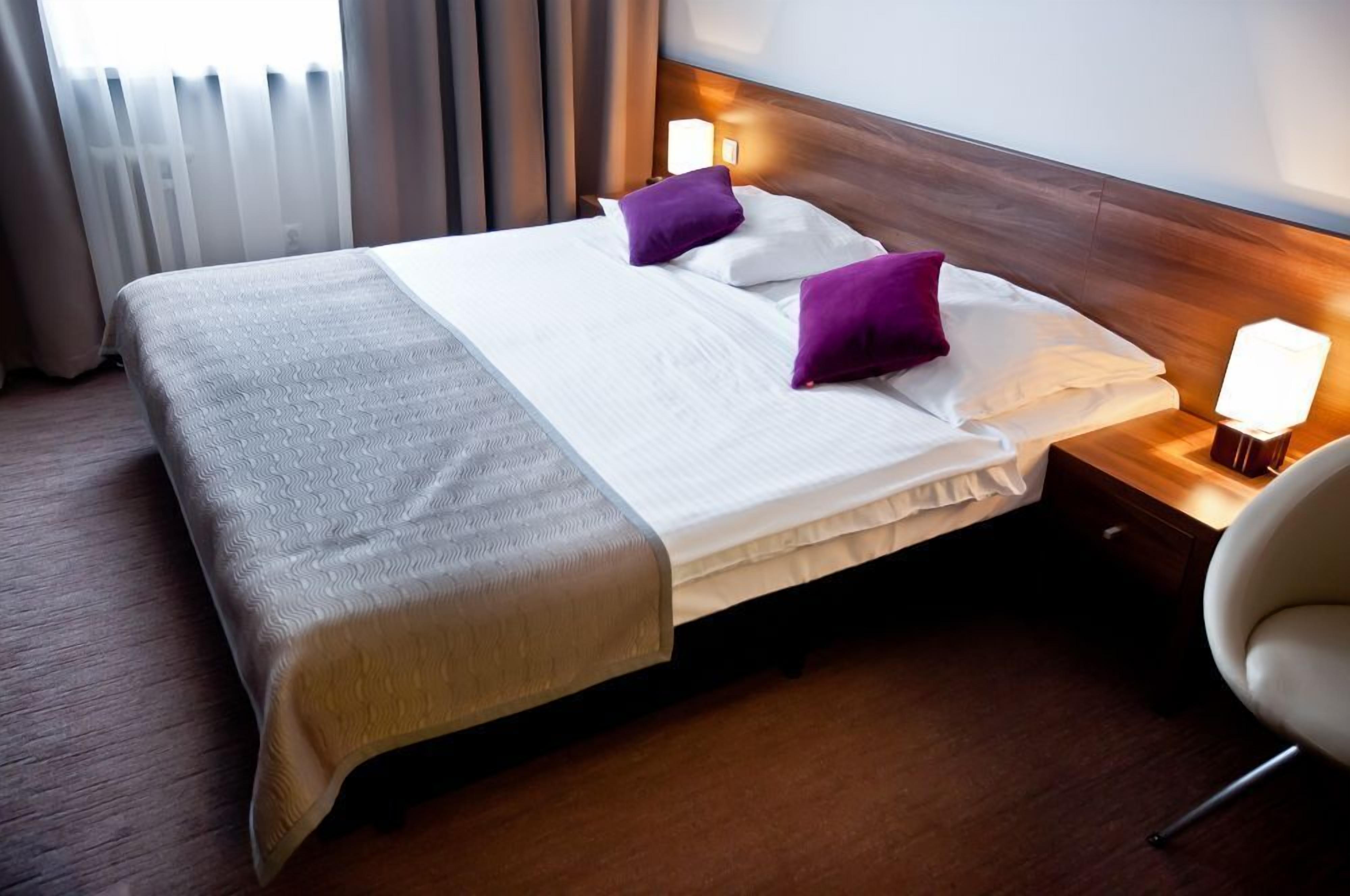 A neatly made bed with a wooden headboard, two purple pillows, and a bedside lamp.