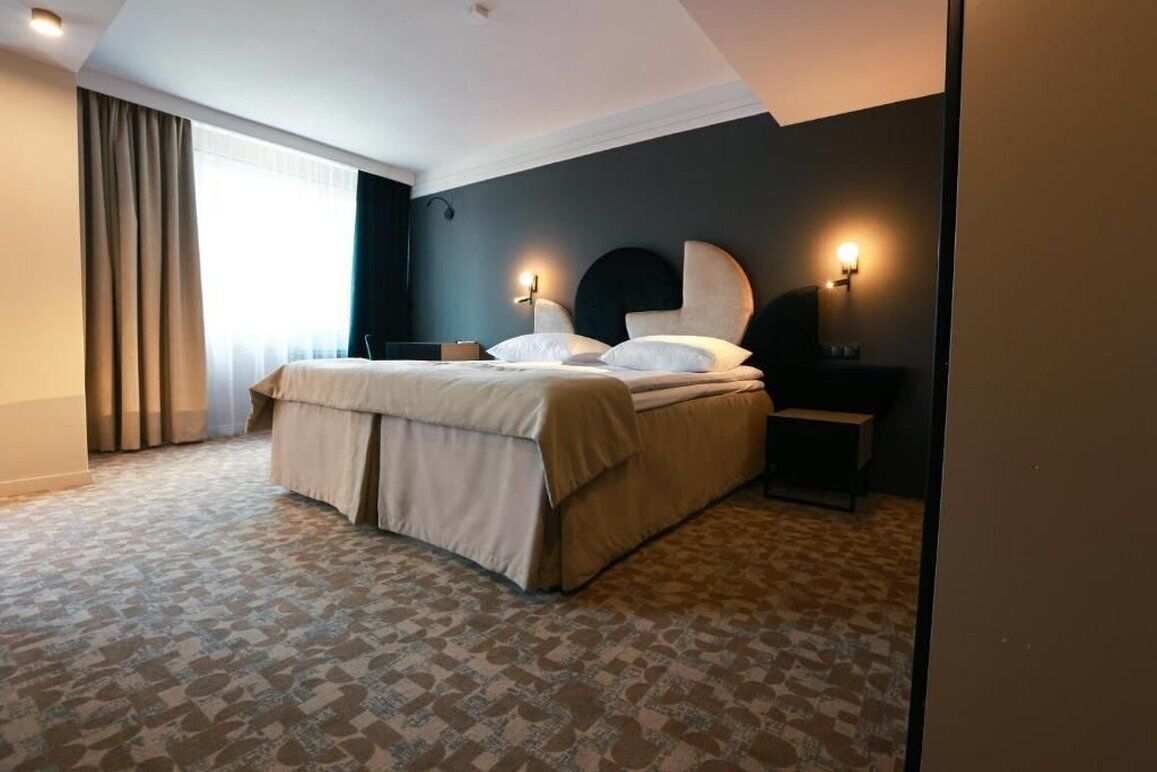 Una habitación de hotel con cama, mesita de noche, lámpara y un amplio ventanal con cortinas.