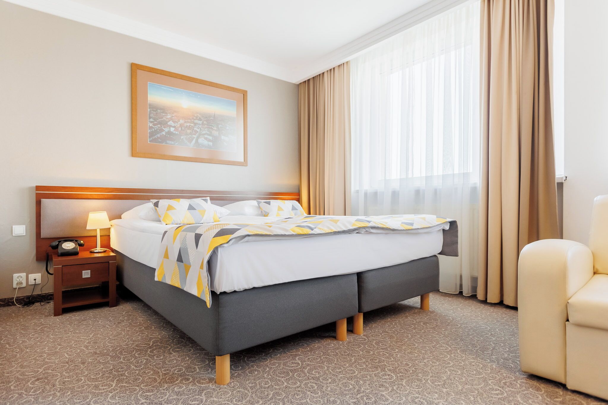 Une chambre d’hôtel avec un lit, une table de chevet avec une lampe, une chaise, une fenêtre avec des rideaux et un tableau encadré au mur.