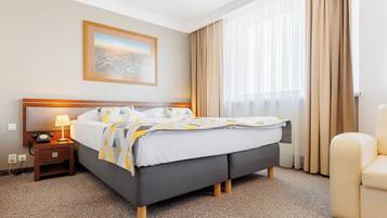 Une chambre d’hôtel avec un lit, une table de chevet avec une lampe, une chaise, une fenêtre avec des rideaux et un tableau encadré au mur.
