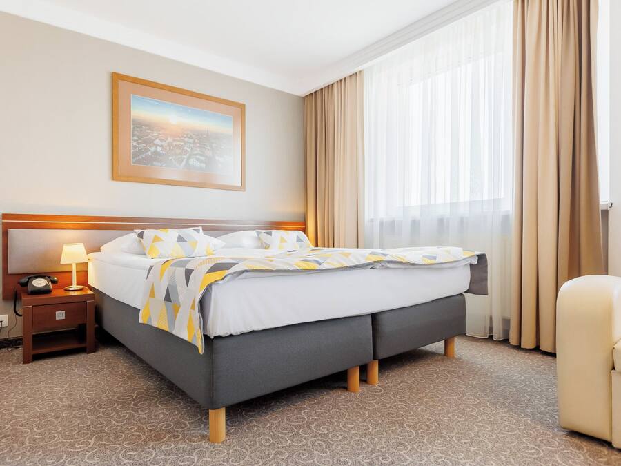 Une chambre d’hôtel avec un lit, une table de chevet avec une lampe, une chaise, une fenêtre avec des rideaux et un tableau encadré au mur.