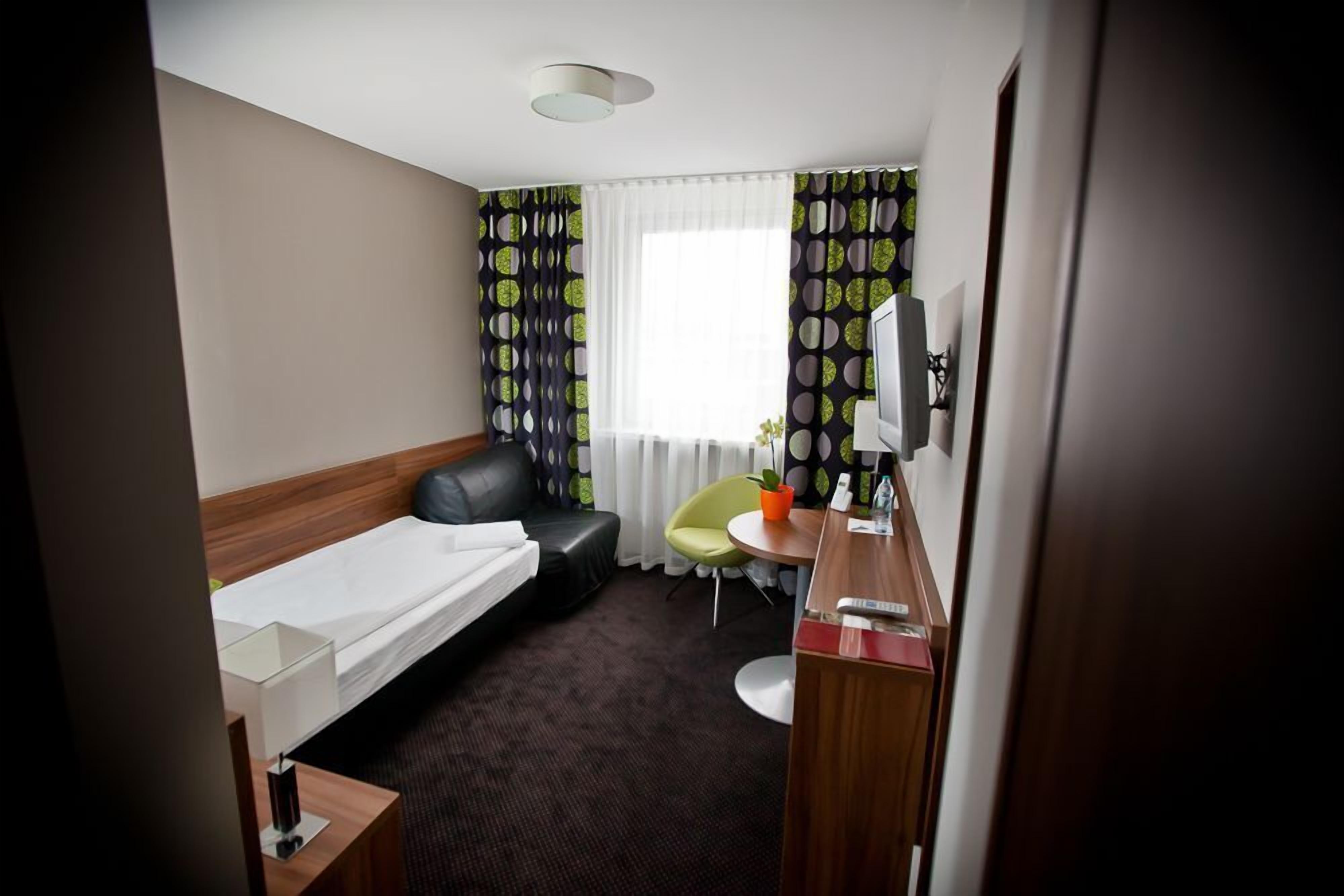 A hotel room with a single bed, a desk with a chair, a small table with a potted plant, and a window with patterned curtains.