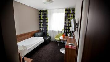 A hotel room with a single bed, a desk with a chair, a small table with a potted plant, and a window with patterned curtains.